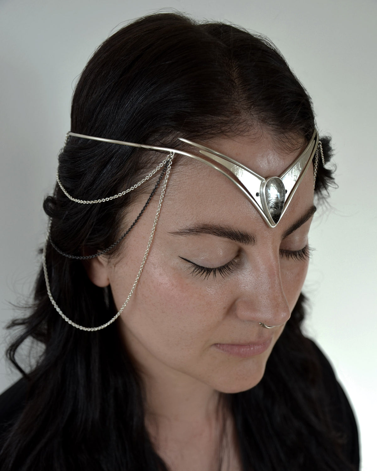 A close-up of a woman with dark hair wearing a silver metal headpiece with delicate chain drapes, photographed against a plain light background. The headpiece is the Odysessy Diadem by Emily Eliza Arlotte.