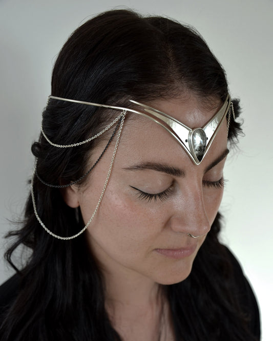 A close-up of a woman with dark hair wearing a silver metal headpiece with delicate chain drapes, photographed against a plain light background. The headpiece is the Odysessy Diadem by Emily Eliza Arlotte.