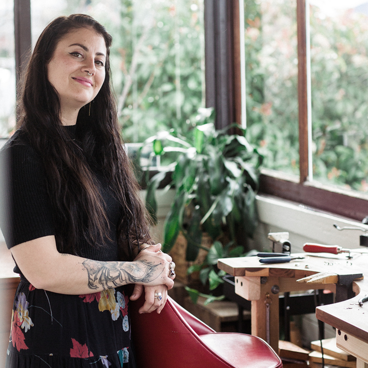 Emily Eliza Arlotte  stands in their bright jewellery workshop studio in Hobart, Tasmania, leaning on a red chair near large windows with greenery outside. They are wearing a black short-sleeve top and a dark floral skirt.