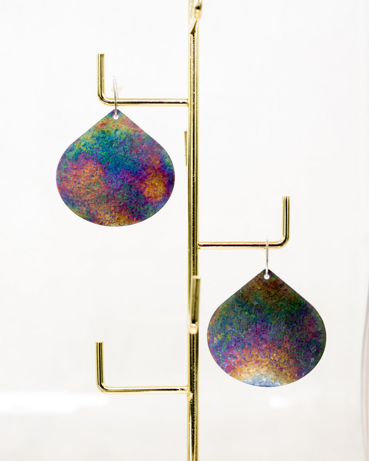 A pair of teardrop-shaped dangling earrings with a colorful, iridescent glitter finish, displayed on a gold metal stand against a white background. The earrings are handmade by Tanja von Behrens, contemporary jeweller.