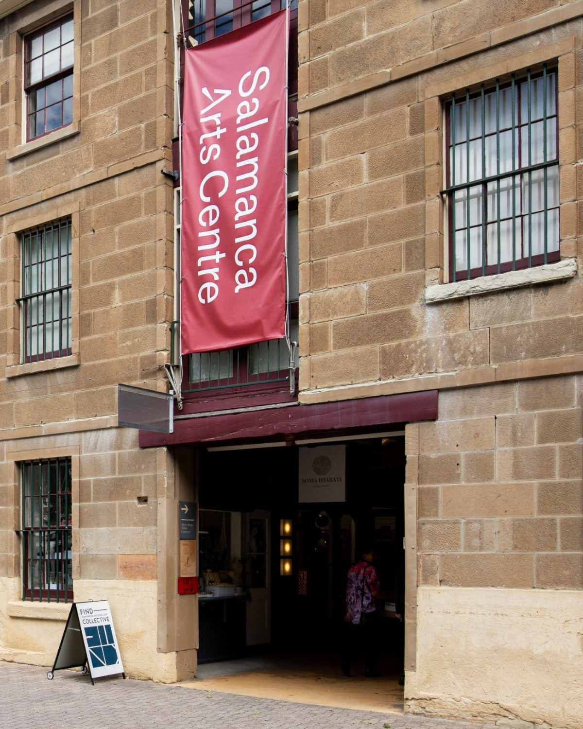 The exterior of the Salamanca Arts Centre building, featuring a warm tan stone facade, barred windows, a prominent red vertical banner hanging from the upper right side and the FIND Contemporary Jewellery Collective signage.