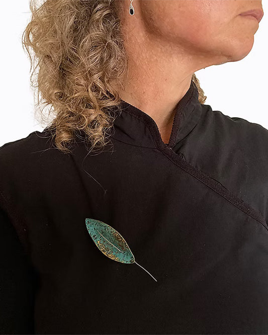 A close-up of a person wearing a black high-neck top, featuring a decorative leaf-shaped brooch in a distressed teal and gold finish pinned to the chest. Handmade Float Pin by Karin Beaumont, in Hobart Tasmania.