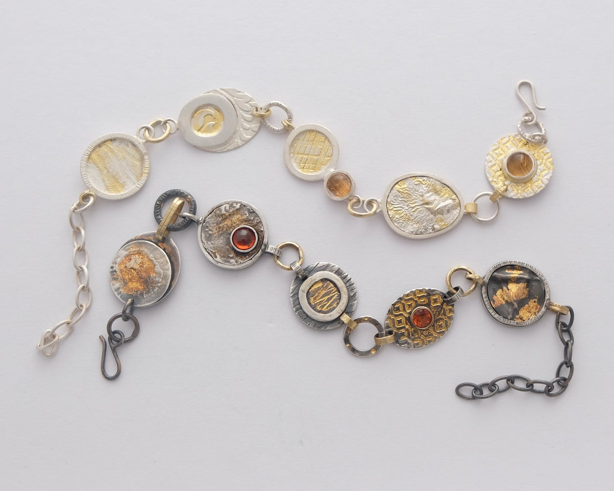A vintage-style mixed metal chain bracelet featuring circular medallions in silver, gold, and bronze tones, some with textured patterns and others containing amber-colored inlays, displayed on a clean white background. Handmade by Megan Dickens in Tasmania, Australia.